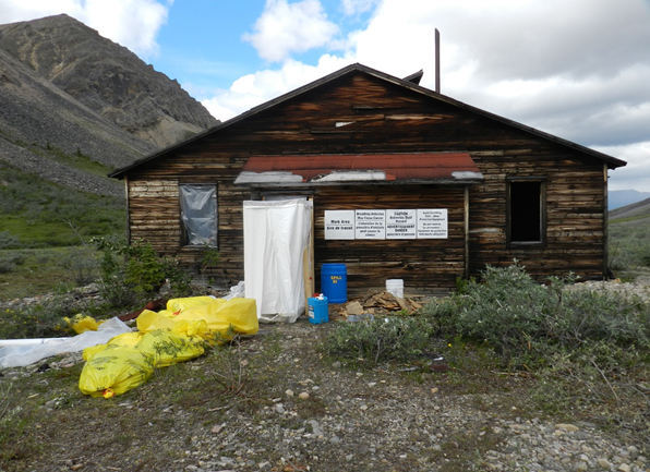 Un vieux bâtiment en bois le long du sentier Canol. Des bâches blanches délimitent une zone qui a été décontaminée et des sacs jaunes de déchets dangereux sont empilés devant le bâtiment. Des montagnes sont à l'arrière-plan.