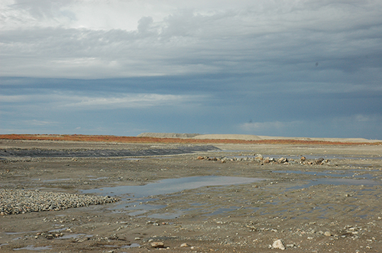 This photo shows a flat area of rock and soil, separated by trees. Forest burnt by the 2014 forest fires is visible behind the structures.