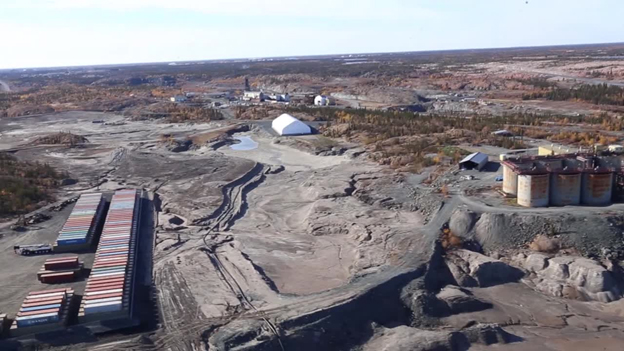 This picture shows a tailings area of crushed rock, with sea cans and old buildings surrounding it.