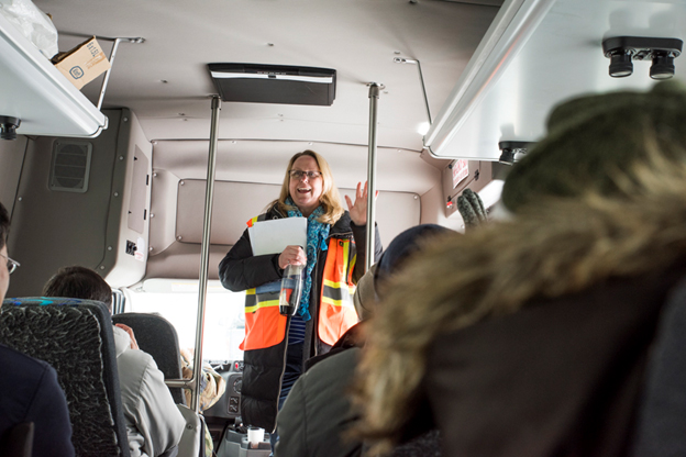 This picture shows a woman in personal protective equipment in a tour bus with other people in personal protective equipment.