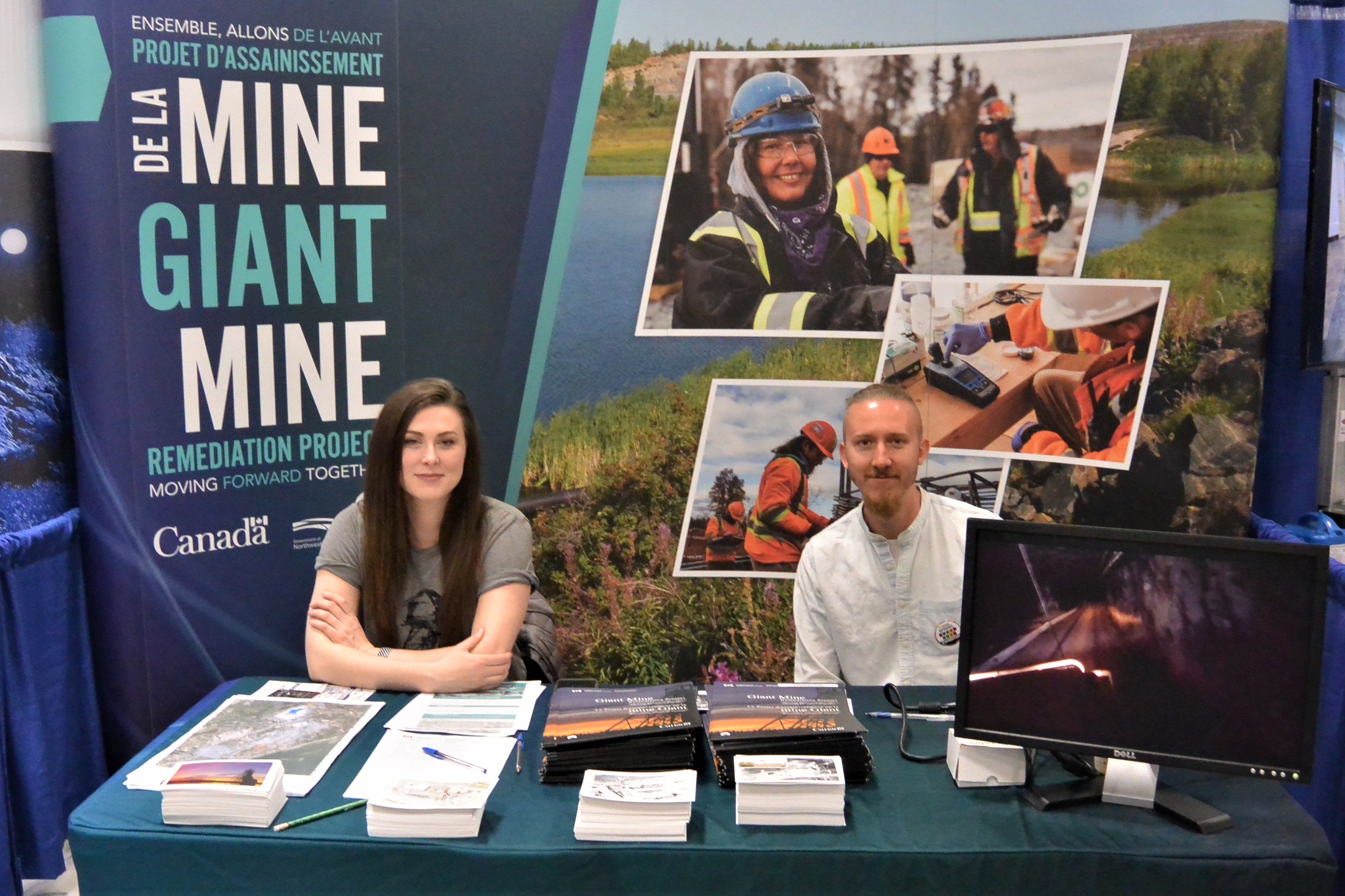 Cette photo montre une femme et un homme assis à la table d'un kiosque d'exposition, avec comme toile de fond l'affiche du projet d'assainissement de la mine Giant.