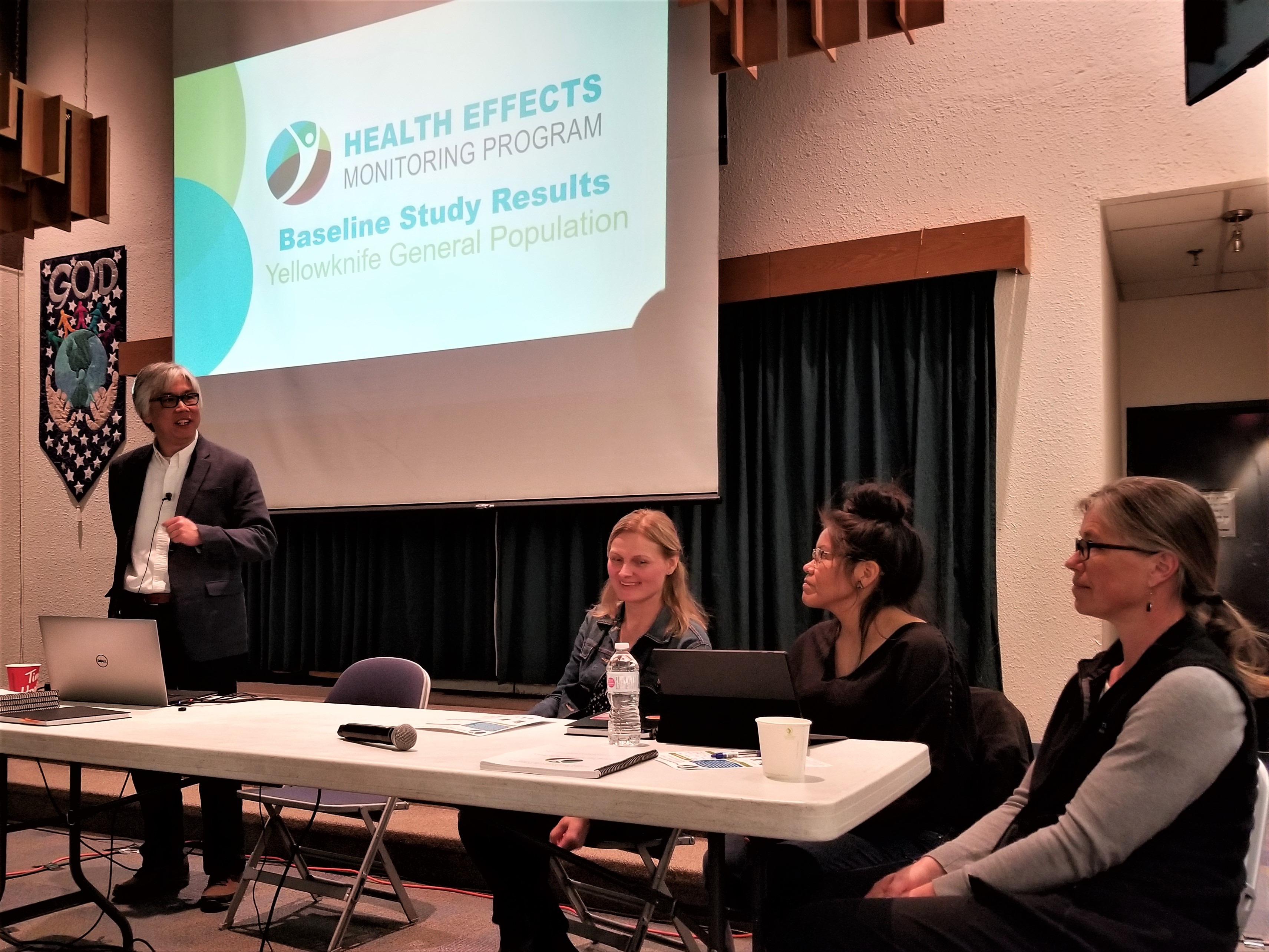 Cette photo montre un homme qui fait une présentation, debout derrière une table. Trois femmes sont assises à sa droite, à la table. Derrière eux, à l'écran, on peut voir une diapositive sur laquelle il est indiqué : « Health Effects Monitoring Program – Baseline Study Results – Yellowknife General Population » (Programme de suivi des effets sur la santé – Résultats de l'étude de base – Population générale de Yellowknife).