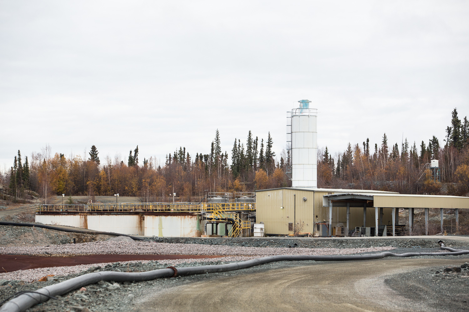 This picture shows a pipe leading up a road to an effluent treatment plant. Three large tanks are visible next a building, with trees in the background.