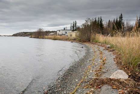 This picture shows a lake shore with pebbled shoreline and natural grass. Old buildings in disrepair are visible along the shore.